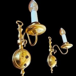 May include: Three antique brass wall sconces with candle-shaped light bulbs. The sconces have a round base with a switch and a decorative arm holding the bulb. The sconces are against a black background.
