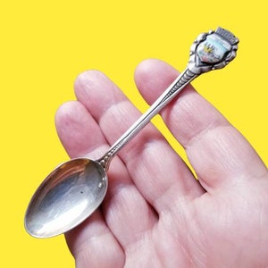 May include: Silver souvenir spoon with a colorful enamel design of a castle on the handle. The spoon is in a hand.