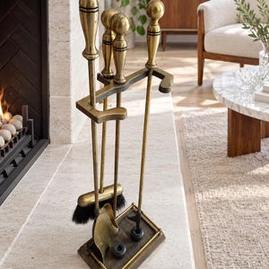 May include: A brass fireplace tool set featuring a stand with a rectangular base, a shovel, a poker, and a brush with black bristles. Each tool has a long handle topped with a spherical finial. The set is constructed from metallic brass.
