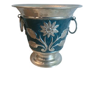 May include: Silver-toned metal vase with a teal-colored band featuring a floral design. The vase has a flared rim, two handles, and a pedestal base. The floral design includes a central flower and leaf accents.
