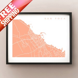 May include: Framed map print of San Foca in coral and white. The map shows the city's streets and coastline. The words "SAN FOCA" are printed in a simple font at the top. The print is in a black frame.