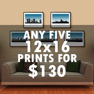 May include: Five framed 30x41 cm prints of city skylines hang above a sofa with green and patterned cushions. The prints feature the cities of Winnipeg, Edmonton, Montreal, and Hamilton. Two small potted trees flank the sofa.