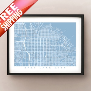 May include: A framed map of Salt Lake City, Utah, in a minimalist style. The city's street grid and surrounding areas are depicted in white against a light blue background. The map is enclosed in a black frame.