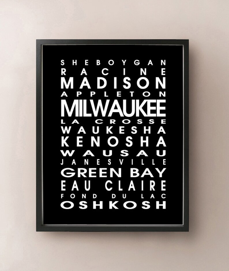 May include: Black framed print featuring a list of Wisconsin city names in white, including Milwaukee, Madison, and Green Bay. The text is arranged vertically on a black background.