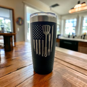 May include: Black insulated tumbler with a silver lid. The tumbler features a laser-etched design of the American flag with grilling tools. The design includes a spatula, tongs, and a fork. The tumbler is designed to hold approximately 20 ounces.