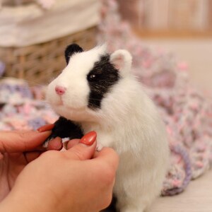 May include: A white and black needle felted hamster with a pink nose and black eyes. The hamster is being held by a person's hands.