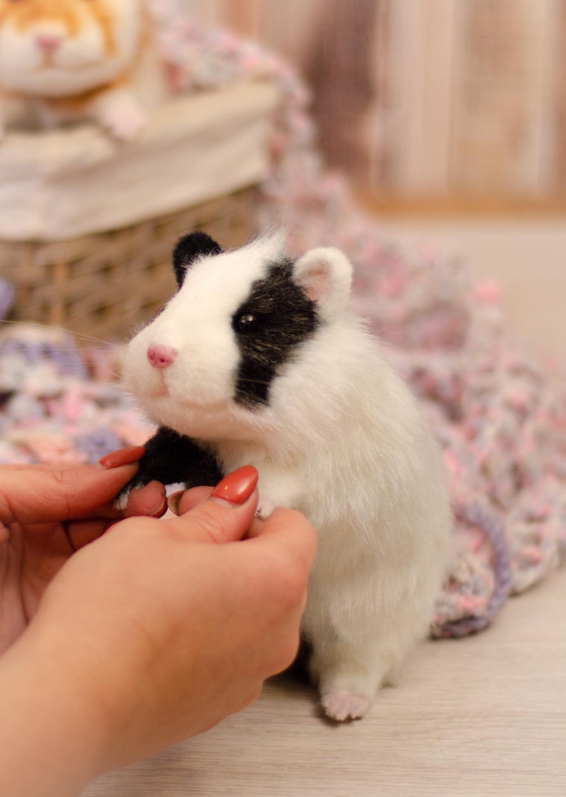 May include: A white and black needle felted hamster with a pink nose and black eyes. The hamster is being held by a person's hands.