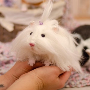 May include: A white fluffy stuffed animal with a purple bow on its head. The animal is being held in a person's hands.