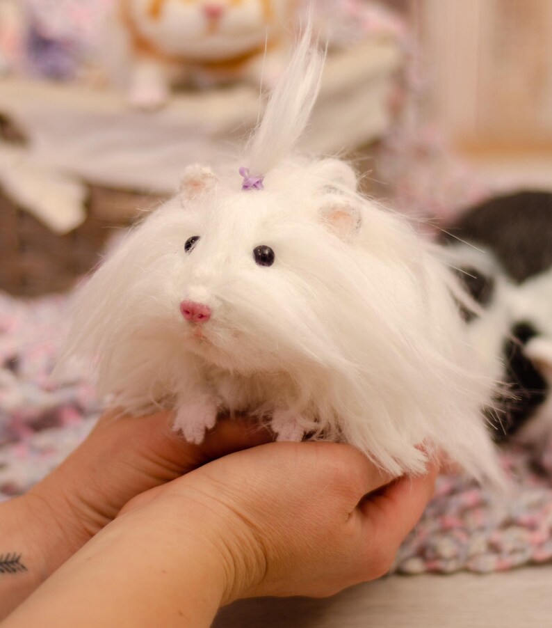 May include: A white fluffy stuffed animal with a purple bow on its head. The animal is being held in a person's hands.