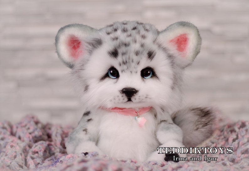May include: A white and gray plush toy with black spots, blue eyes, and pink ears. The toy is wearing a pink collar with a charm. The toy is sitting on a pink and gray blanket. TEDDIKTOYS Irina and Igor.