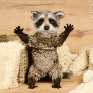 May include: A handcrafted raccoon plush toy with a realistic design. The plush has a gray and brown fur coat, black paws, and a black mask around its eyes. It is standing with its arms raised, near a woven basket and a cream-colored blanket.