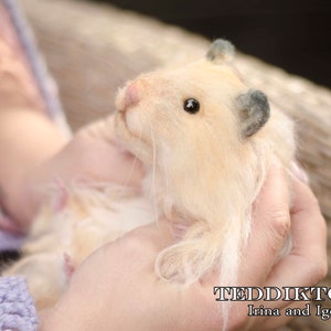 May include: A close-up of a light brown, fluffy, handmade hamster toy with black eyes and a gray ear. The toy is being held by a person's hands.