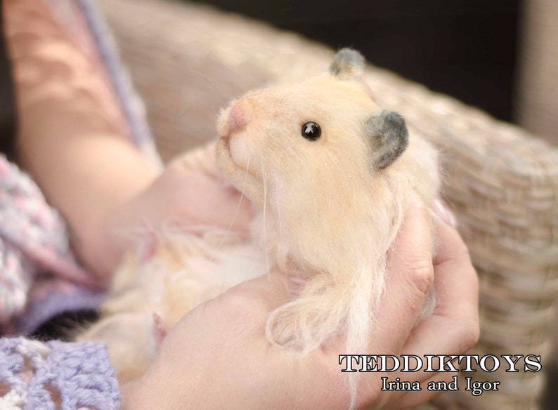 May include: A close-up of a light brown, fluffy, handmade hamster toy with black eyes and a gray ear. The toy is being held by a person's hands.