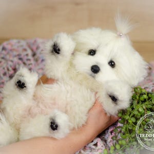 May include: A fluffy, white stuffed dog toy with black paw pads and a small ponytail. The toy is lying on its back, held by a person. The background includes a patterned blanket and greenery.