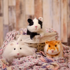 May include: Three stuffed hamsters in a wicker basket on a pink and purple crocheted blanket. One hamster is black and white, one is white with a pink bow, and one is orange.