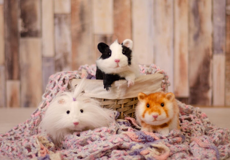 May include: Three stuffed hamsters in a wicker basket on a pink and purple crocheted blanket. One hamster is black and white, one is white with a pink bow, and one is orange.