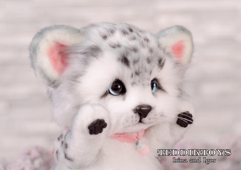 May include: A white and gray plush toy with black spots, blue eyes, and pink ears. The toy is wearing a pink ribbon with a cupcake charm. The toy is called "Teddiktoys Irina and Igor".