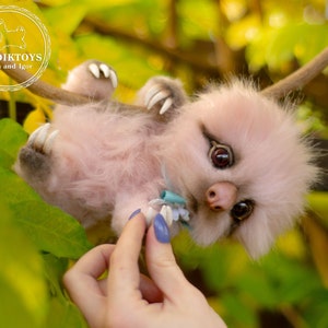 Puede incluir: Una criatura peluda rosa y gris con ojos grandes y un hocico pequeño cuelga boca abajo de una rama. La criatura lleva un collar en forma de flor azul y blanco. La criatura está siendo sostenida por una mano con uñas pintadas.