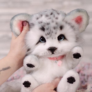 May include: A white and gray plush toy snow leopard with blue eyes and black paw pads. The toy is wearing a pink collar with a cupcake charm. The toy is being held by a person's hands.
