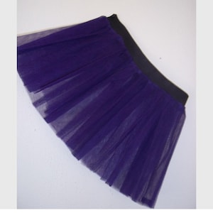Pode incluir: Uma saia tutu curta e em camadas em um tom roxo profundo. A saia apresenta várias camadas de tecido transparente, criando uma silhueta cheia e volumosa. Possui uma cintura elástica preta larga para um ajuste confortável. A saia é projetada para dança ou fantasias.