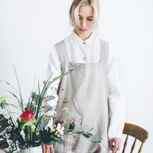 May include: A woman wearing a light grey linen apron stands at a table set with a pink tablecloth. The table is decorated with a vase of flowers, a cup, a plate of butter, a glass of coffee, and a plate of biscuits.