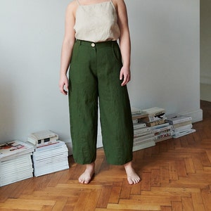 May include: A woman wearing a light beige tank top and dark green wide-leg trousers. She is standing on a wooden floor with stacks of books and magazines behind her.