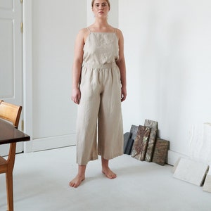 May include: A woman wearing a beige linen jumpsuit with a square neckline and wide legs. She is standing in a white room with a wooden table and some decorative tiles on the floor.