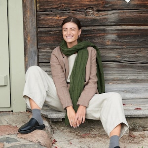 May include: A woman wearing a tan blazer, cream-colored pants, and black shoes sits on a wooden porch. She is wearing a green scarf. The background features a wooden wall with a white diamond-shaped window.