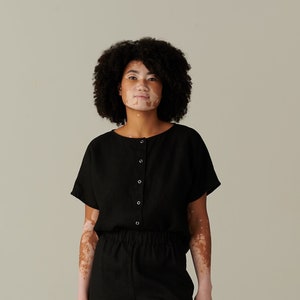 Pu&ograve; includere: Una donna indossa una tuta in lino nero con una parte superiore abbottonata e una vestibilit&agrave; rilassata. Indossa mocassini in camoscio marroni.