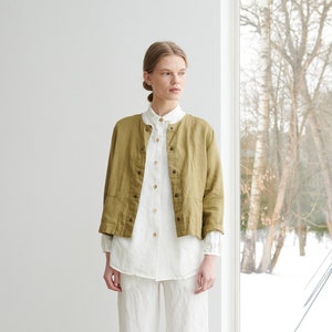 Puede incluir: Una mujer con una camisa de lino blanca, una chaqueta de lino verde oliva, pantalones de lino blancos y sandalias de cuero negras.