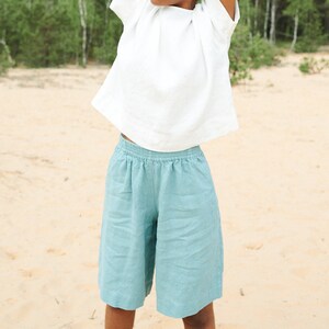 May include: A woman wearing a white linen top and light blue linen shorts stands in a sandy area with trees in the background. She is stretching her arms above her head.