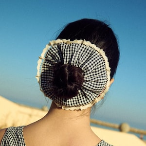 Peut inclure: Un accessoire pour cheveux à carreaux noirs et blancs avec une bordure en dentelle crème est porté en chignon. La personne porte un haut sans manches à carreaux noirs et blancs. Le fond est un ciel bleu clair et une plage.