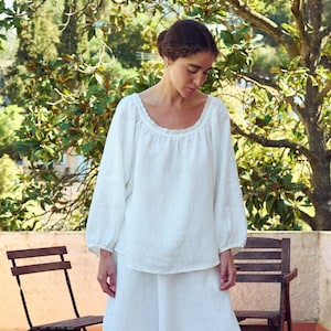 May include: A white linen blouse and skirt set. The blouse has a round neckline with lace trim and long sleeves. The skirt is a wide, flowing style. The outfit is worn with black flats. The background includes trees and a brick patio.
