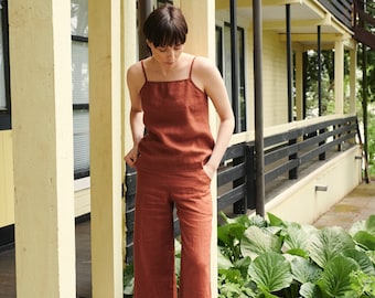 Puede incluir: Una mujer lleva un mono de lino color óxido con silueta de pierna ancha. Está de pie en un patio de concreto frente a un edificio amarillo.