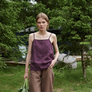 May include: A woman wearing a purple linen tank top and brown linen trousers walks on a gravel path in a woodland setting. She is holding a bunch of green leafy plants.