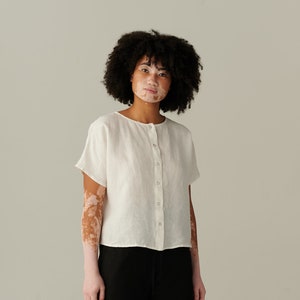 May include: A white linen button-down shirt with short sleeves and a relaxed fit. The model is wearing black pants and black shoes.