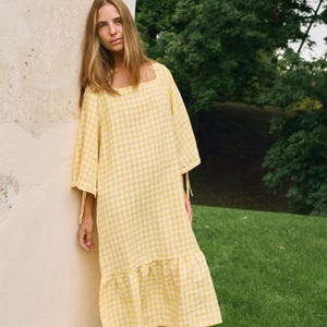 May include: A yellow and white gingham print dress with a square neckline and elbow-length sleeves. The dress has a ruffled hem and is knee-length. The model is leaning against a beige wall, with a green background.
