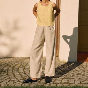 May include: A person wearing a yellow linen tank top and beige linen pants. The outfit is paired with black slip-on shoes. The person is standing on a cobblestone walkway in front of a white building.