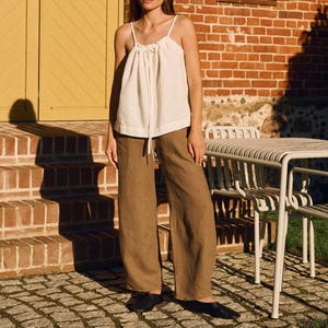 Può includere: Una persona indossa un top bianco con spalline sottili e pantaloni di lino marroni. L'outfit è abbinato a ballerine nere. Lo sfondo presenta un muro di mattoni, una porta gialla e un tavolo in metallo bianco.