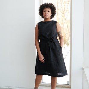 May include: A woman wearing a black linen dress with a tie belt. She is standing in front of a window with a white frame. She is wearing black shoes and socks.