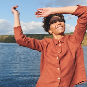 May include: A person wearing a rust-colored linen shirt and matching pants, standing in front of a body of water. The person is smiling and has their arms raised.