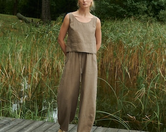 May include: A woman wearing a light brown linen tank top and matching wide-leg pants stands on a wooden dock. She is in a natural setting with tall green grass and trees in the background.