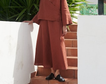 May include: A woman wearing a rust-colored linen wrap top and skirt. The top has a V-neck and long sleeves. The skirt is a midi length and has a wrap style. She is wearing black leather sandals.