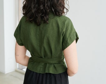 May include: A woman wearing a green linen wrap top with a black linen skirt. The top has short sleeves and a tie at the waist.