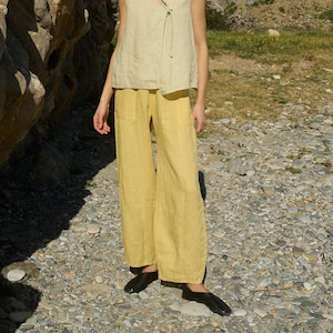 Può includere: Un top senza maniche giallo chiaro con scollo a V e chiusura con fiocco, abbinato a pantaloni ampi giallo senape. L'outfit è completato da ballerine nere. L'abbigliamento sembra essere realizzato in lino o in un tessuto leggero simile, adatto alla stagione calda.