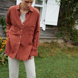 May include: A woman wearing a rust-colored linen shirt with a button-down front and a chest pocket. She is also wearing light gray linen pants.
