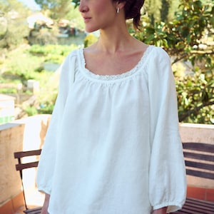 May include: White linen blouse with a square neckline and lace trim. The long sleeves gather at the wrist. The blouse is part of a two-piece set. The model is standing outside on a sunny day.