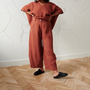 May include: A woman wearing a rust-colored linen top and pants. The top has a wide, loose fit and a V-neck. The pants are wide-legged and have a high waist. She is wearing black leather slip-on shoes.