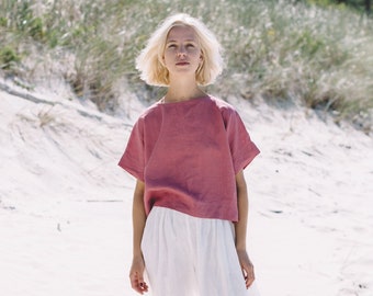 May include: A woman wearing a pink linen top and white linen pants stands on a sandy beach. She is looking up at the sky.