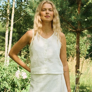 May include: A woman wearing a white linen outfit, consisting of a button-down tank top and a wrap skirt. The outfit is set against a backdrop of lush greenery, including trees and flowers, creating a natural aesthetic.